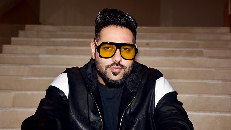 Badshah looking at the camera wearing a black leather jacket with white shoulder pattern. Hee's wearing yellow tinted glasses with his hair slicked up. The background is of light brown coloured long steps.