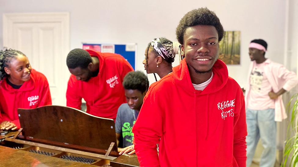 Fredlin Morency from the Reggae Roots choir wearing a red hoodie, smiling at the camera with members of the choir facing away in the background.