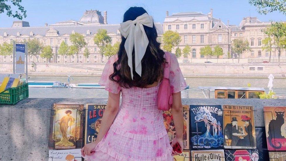 A girl is facing away from the camera, looking out onto a city scene with a boat on a river and grand buildings in the background. She is wearing a pink and white gingham dress with pink flowers printed across it. The sleeves of the dress are made of a floaty, chiffon material. Her hair is in a half up half down hairstyle with a big white bow holding up the ponytail part of it. She has a hot pink bag on her right shoulder and is carrying a phone with a red case.