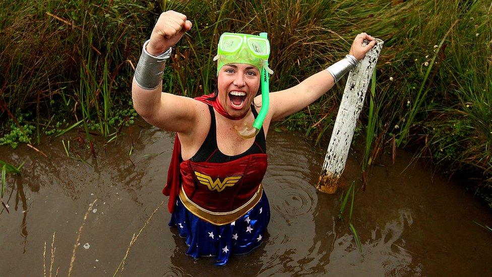 A person wearing a superhero costume cheers in celebration whilst stood in a muddy peat bog.