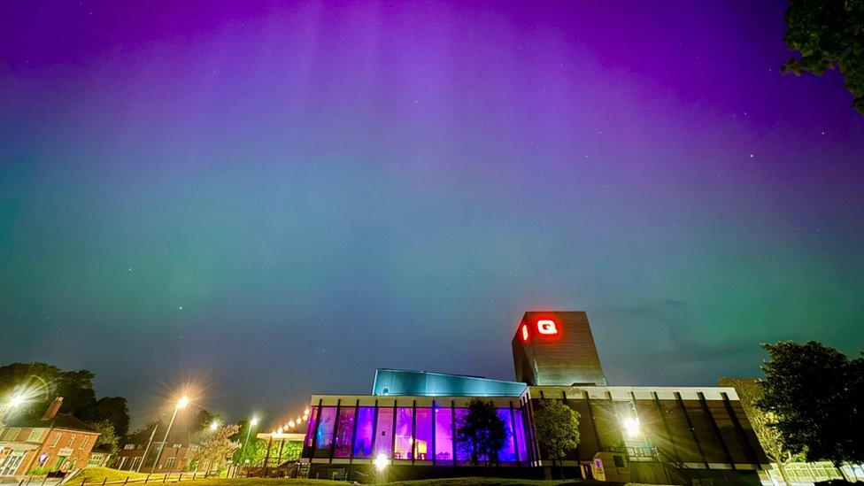 Green, blue and pink lights in the sky behind the Queen's Theatre building, lit up by its own purple and blue lights