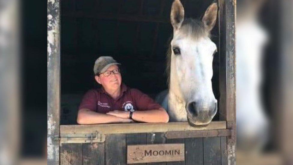 A woman in a burgundy top and a cap standing next to a white stallion