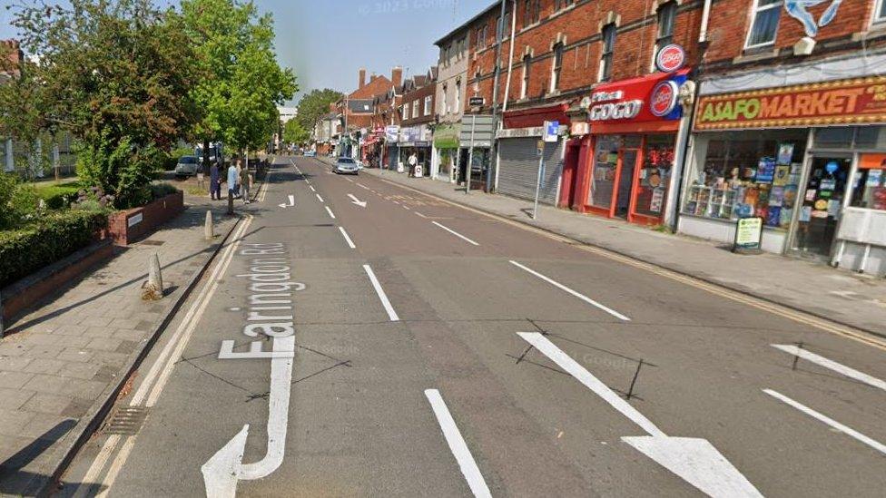 A shot of Faringdon Road with pavements on both sides, shops to the right and greenery to the left