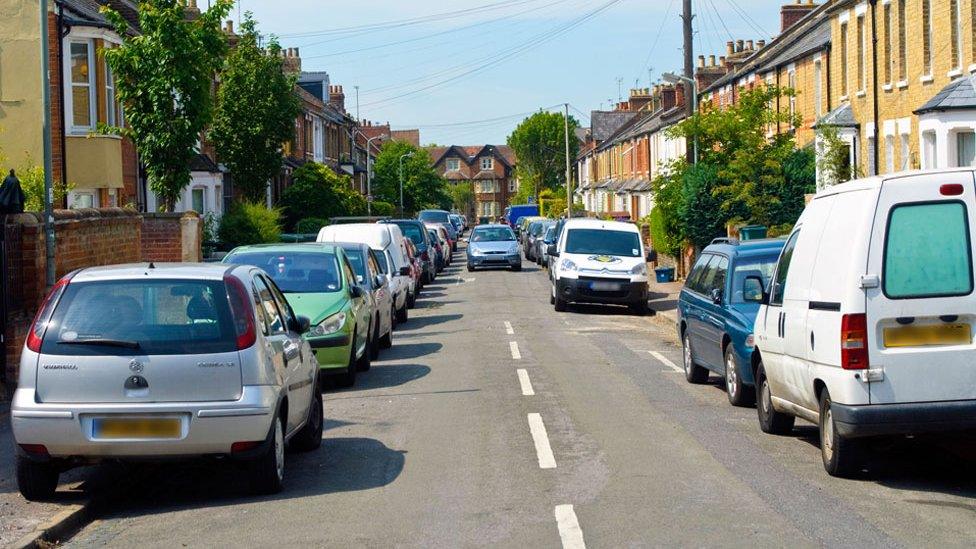 Cars parked on both sides of road, on pavements, with a car coming down the road