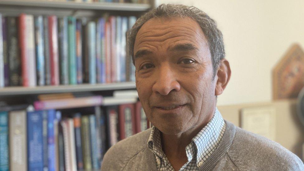 Dr Lee Riley stands looking at the camera, in front of a heavily-stocked bookcase