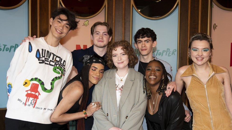 A group shot featuring seven people, they're all young. In the centre is a female, wearing a pale, greyish jacket over a white t-shirt. Her hair is red and styled in ringlets. A young black female wearing a black bandana with paisley pattern, and a sleeveless top, is leaning on her right shoulder. Leaning on her other shoulder, another young black female wearing a black leather jacket, her hair styled in locs, smile-laughs towards the viewer. Next to her, a young, white, woman with short dark hair, highlighted blonde, wears exaggerated blue eyeshadow and a gold sleeveless dress with oversized collar. Behind the woman in the centre, on her left, a young white man wearing a powder blue suit jacket/blazer over a black t-shirt can be seen. He's got short, dark, curly hair. Next to him is a young, white man dressed in black, his hair is centre-parted into curtains. Finally, on the far-left, a young man who's much taller than the others leans into shot. He's wearing a white sweatshirt.