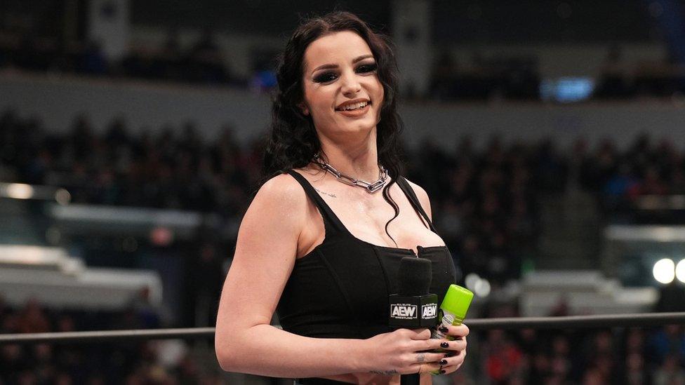 Saraya smiles at the camera. She's in the middle of a wrestling ring, holding a microphone and wearing a vest top and chunky chain necklace