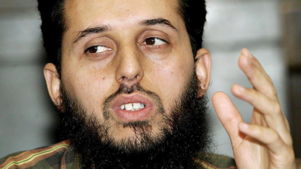 A bearded man gestures with his left hand, looking at something off-camera, in this close-up photo