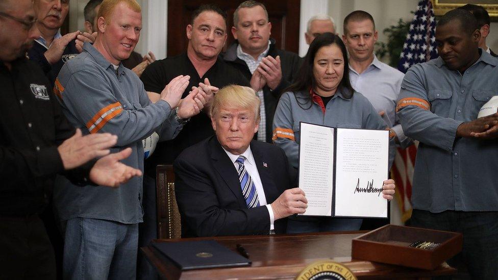 President Donald Trump holds up a piece of paper with his signature while surrounded by steel workers in uniform