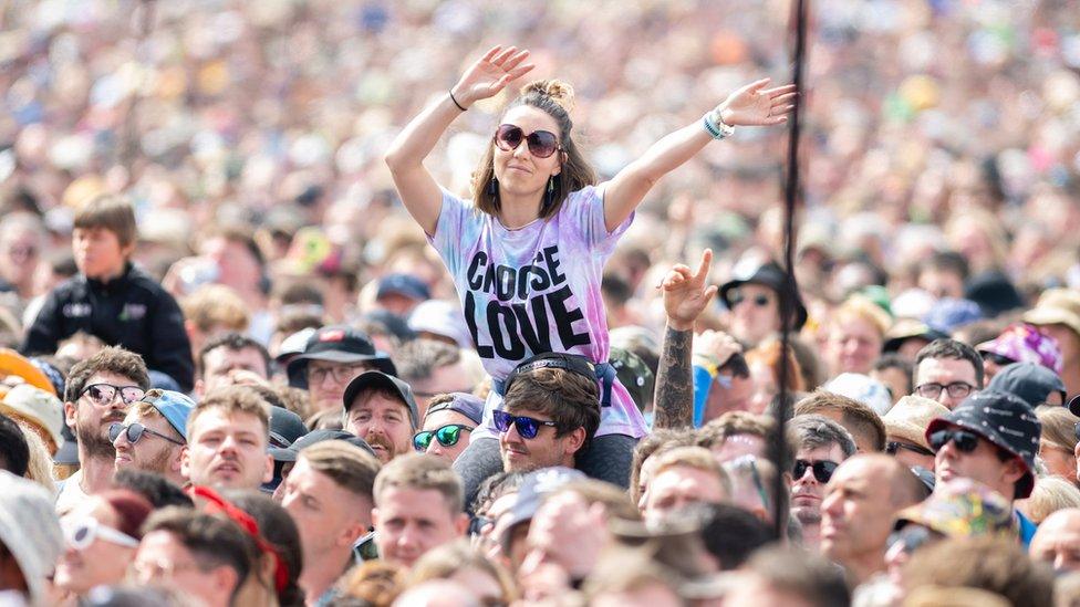 a woman wearing a t-shirt that says choose love while sitting on a friend's shoulders in a massive crowd of people watching performers at glastonbury