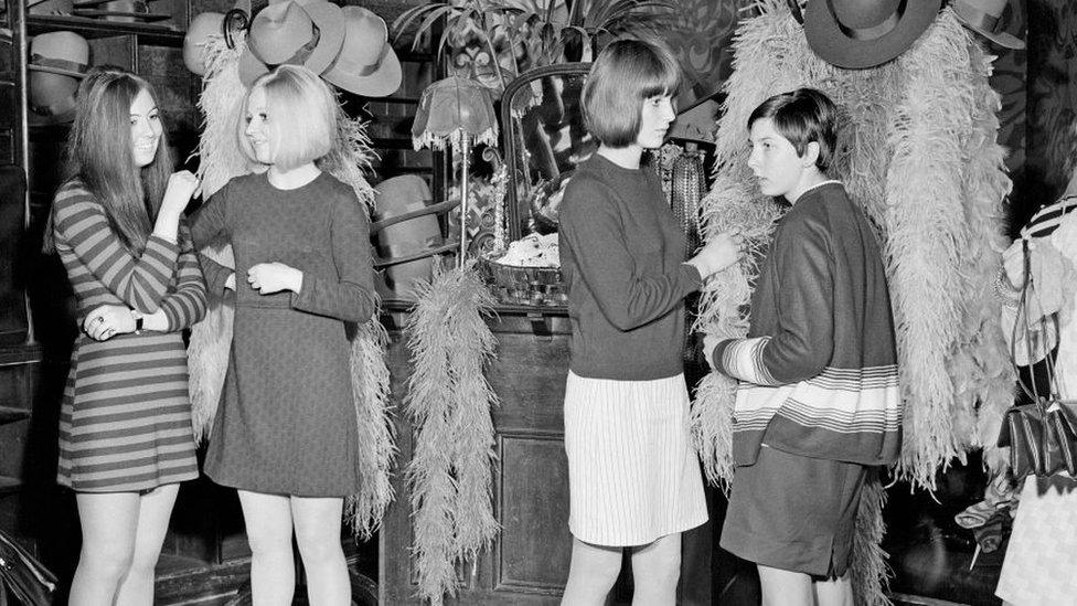 Young women talk together in front of racks of clothes, hats and scarves at the Biba store