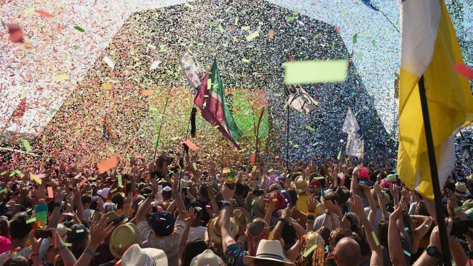 The Pyramid stage at Glastonbury, there is a crowd of people in front of it, with confetti flying everywhere