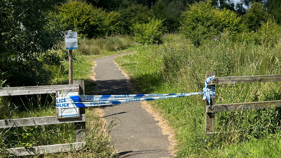 Path through grassy nature area, with fence and police tape in the foreground.