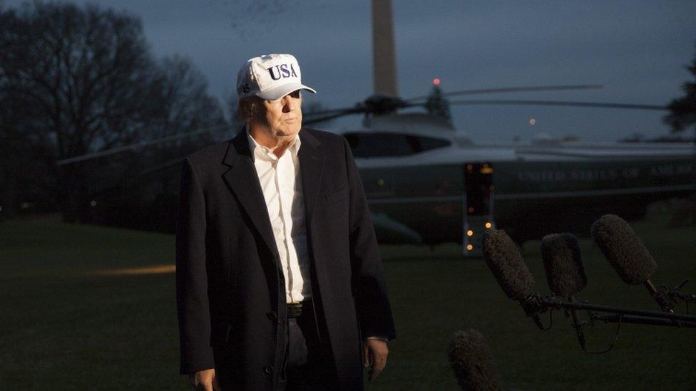 President Trump, wearing a USA cap and white shirt with a a jacket, speaks to media with helicopter in background