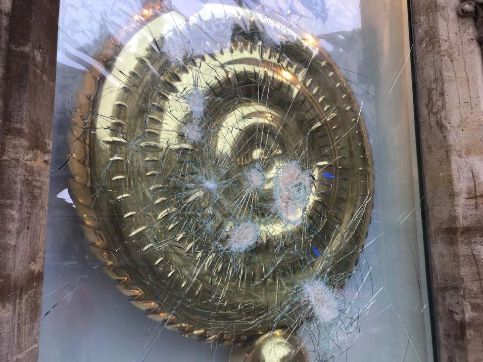 The gold clock is behind glass which has been partially blown out but not entirely smashed. It has large cracks across the glass from the points of impact.