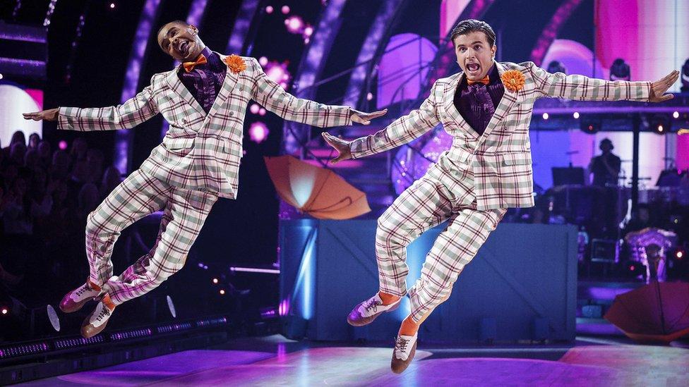 Nikita Kuzmin, Layton Williams during one of their routines. They wear matching cream, grey and burgundy checked suits over purple shirts with orange bow ties, socks and orange flowers tucked in their lapels. They are photographed mid jump, arms outstretched and legs swung to their right, heels together. The stage is set behind them with purple lighting and orange umbrellas