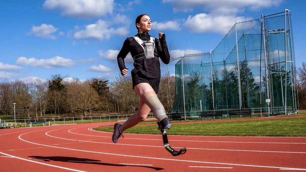 Milly Pickles running on an athletics track. Milly is a white woman in her 20s with brown hair tied back. She is pictured mid-stride, her black running blade extended in front of her. She is wearing a long-sleeved black top, black shorts, and a white trainer as well as a running vest with a water bottle tucked in the front pocket. She has a determined expression and is photographed outside on a blue-sky day. She is running on a red asphalt running track and behind her is a discuss throw and a tree-lined park.