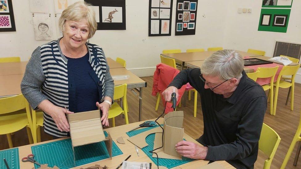 A woman with a striped cardigan and a man with a black jumper, both building cardboard figures