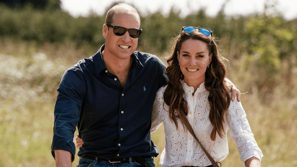 William is wearing sunglasses, jeans and a blue shirt and Kate is wearing a white shirt and jeans. A pair of sunglasses rests on Kate's head.