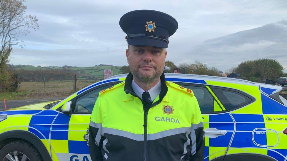 It shows Garda Supt David Kelly in front of a patrol car with fields behind