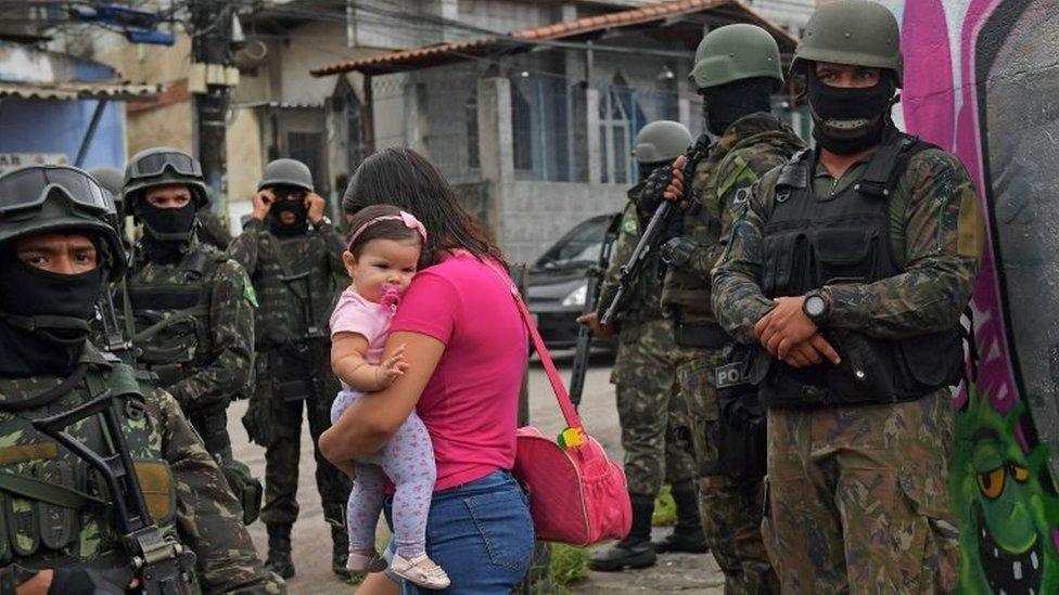 A woman carries her child as she walks past military police on patrol near the Vila Kennedy favela in Rio de Janeiro on February 23, 2018.