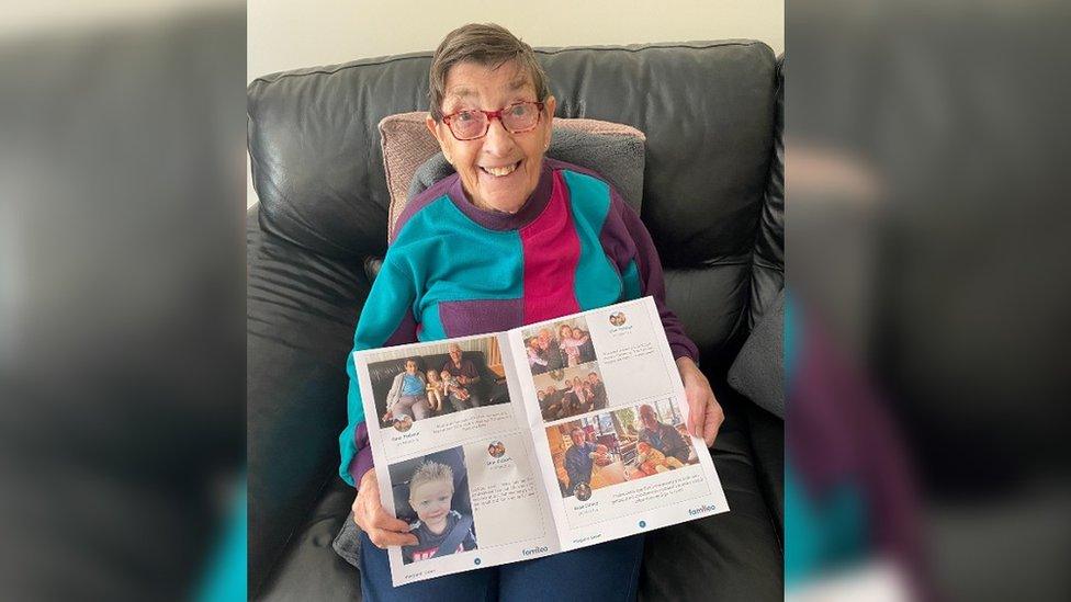 An elderly woman with red glasses and brown hair reading a personalised newspaper sitting on a black couch