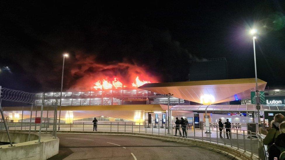 Flames visible on the roof of an airport car park. Some passengers are visible standing on the airport roadway.