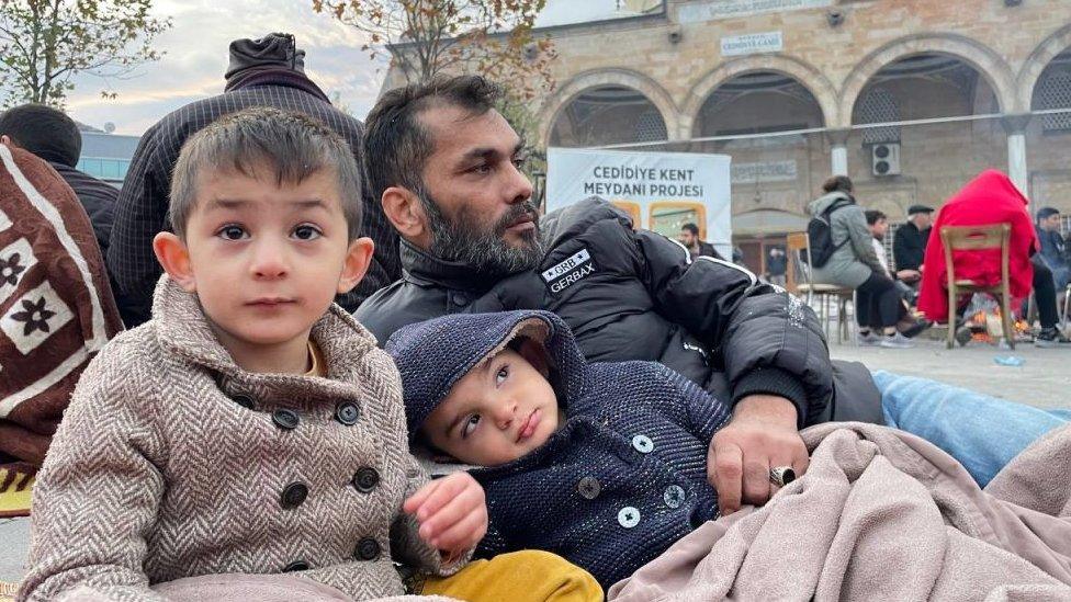 Two children sit in an emergency evacuation centre with their father