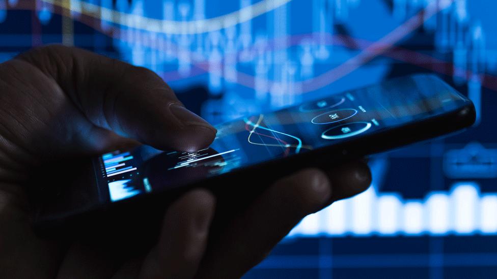 Man holding a phone with financial markets graphs on it