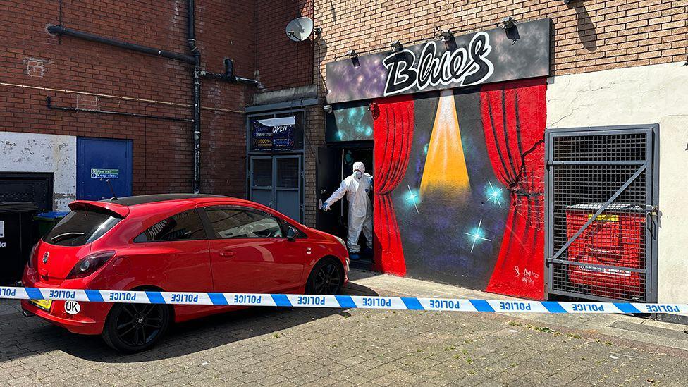 Police tape cordon around a red Corsa car parked outside a bar with the sign in graffiti saying 'Blues'. A CSI police officer wearing white personal protective equipment (PPE) is standing in the entrance holding the front door open by its handle.  