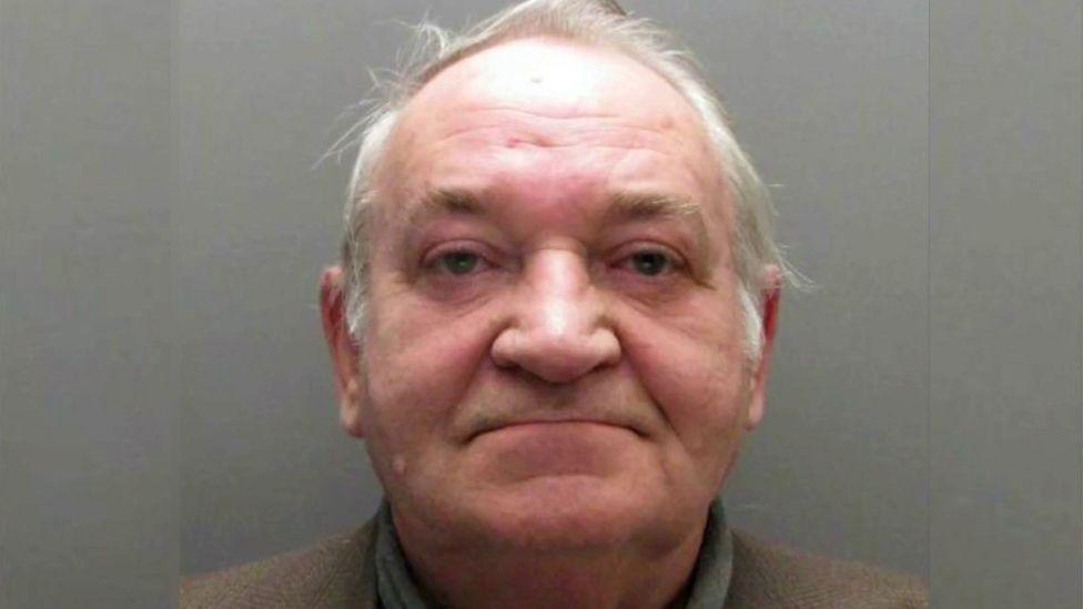 A mugshot of Neville Husband, an older man.  He has short white hair and dark eyes.  He is wearing a turtleneck top and a grey suit jacket. 