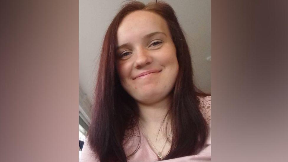 A young woman with long, red hair, smiling at the camera, she is wearing a pink top and gold necklace chain.