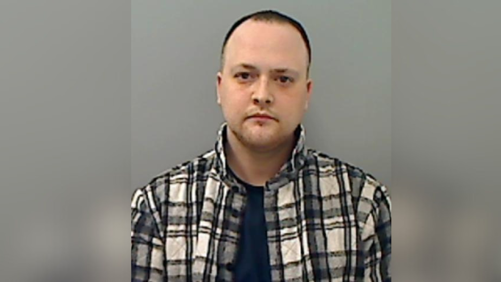 A mugshot of Daniel Dixon who has short black hair, a receding hairline and light facial hair. He is wearing a black and white check shirt unbuttoned with a dark blue t-shirt underneath.