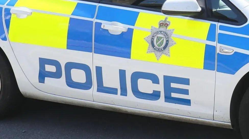 A close-up of the side of a police car. It is white with bright blue and yellow decor. The word "police" is printed in large blue letters. A Lincolnshire Police badge - a star topped with a crown - can be seen on the door.