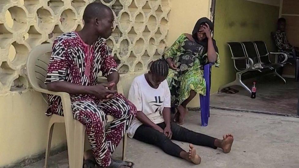 A sreengrab showing three people sitting down sadly outside a hospital. One woman has her head in her hands, another is looking down