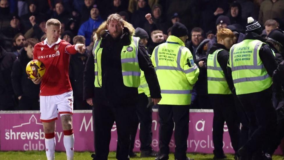 James McClean holding a yellow football, stood on the pitch pointing in front of the crowd. Four stewards in hi-vis gear are seen standing near to him.