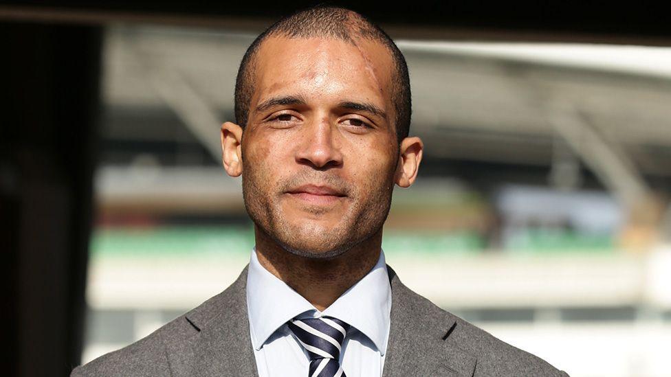 Head and shoulders image of Clarke Carlisle smiling with a sports ground blurred in the background. He is wearing a grey suit jacket, pale grey ad shirt navy and white striped shirt. He has very short dark hair and stubble with scars on his face and head