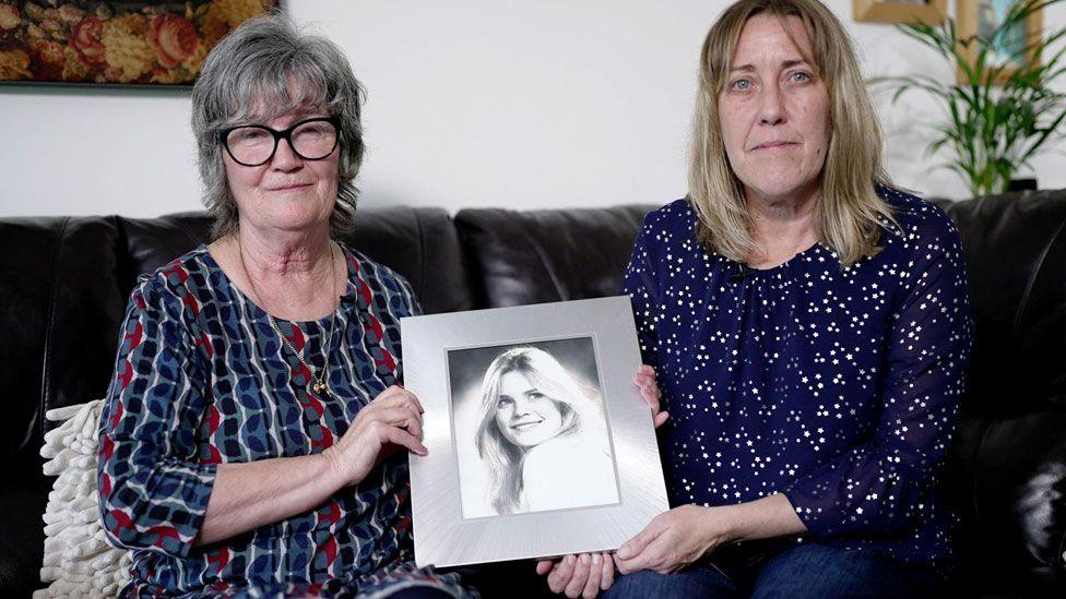 Wendy Ramsay and daughter Emma, sitting on a leather sofa in a living room holding a picture of Samantha. Wendy has grey hair and glasses and wears a blue and red top, while Emma is blonde and wearing jeans with a navy top with a pattern of small stars.