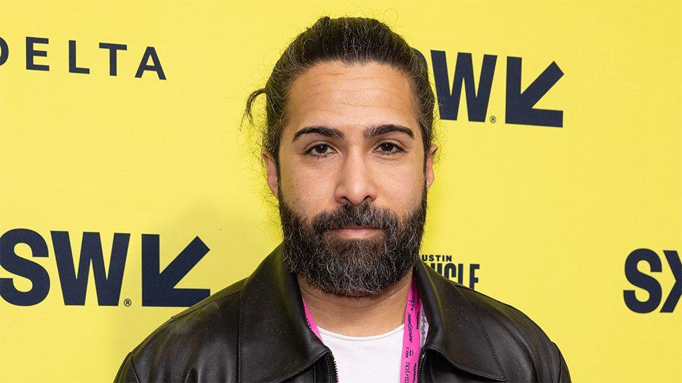 Savan, a man, wearing a black leather jacket, with the background a yellow boarding with the letters SW on it.