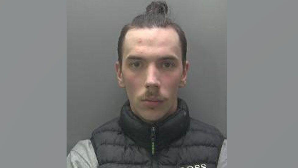 A slightly blurry police mugshot of David Gabryelski, who is clean shaven with a moustache and dark hair pulled into a bun on the top of his head. He is wearing a black puffa waistcoat over a grey top. 