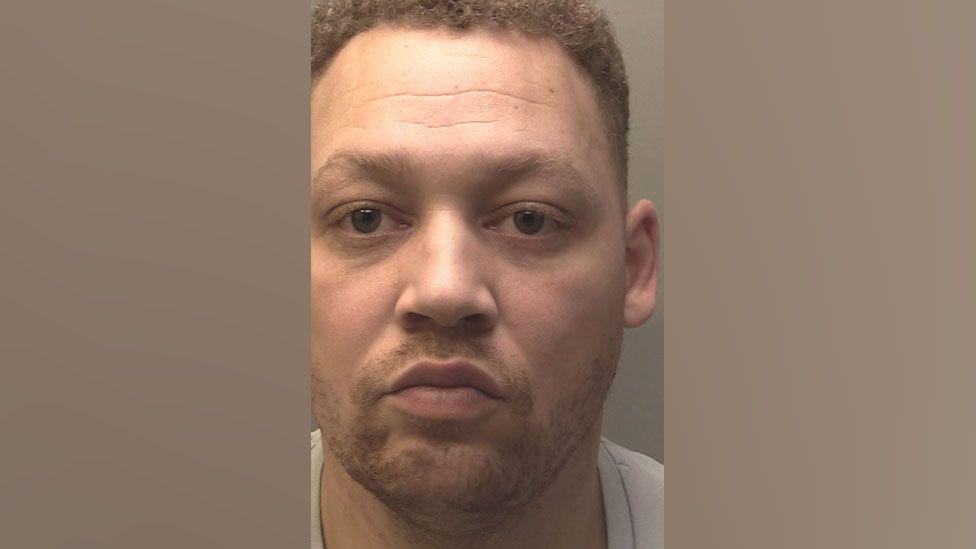 Police custody photograph of Raymond Nicholls who has short brown hair and is wearing a light grey t-shirt