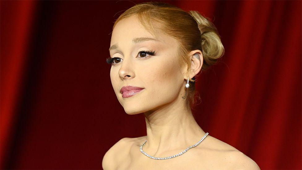 Ariana Grande, a female singer and actress, at a gala. She is wearing a shiny necklace, looking into the distance with a red backdrop.