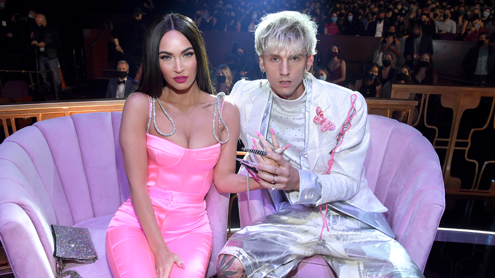 Megan Fox, wearing a pink dress, with silver straps and pink fingernails, holding hands with Machine Gun Kelly, wearing Silver shorts and a silver jacket. The pair are sitting on a purple sofa.