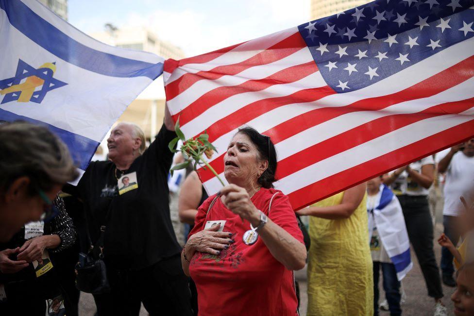 Woman holding flower seems overcome with emotion, as crowds behind wave USA and Israel flags