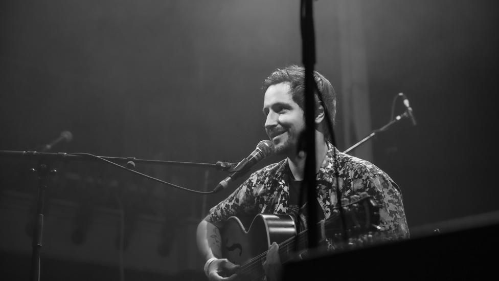 Black and white image of Ollie Thomas sat on stage with his guitar, in front of a microphone, smiling. Ollie has short hair and dark stubble. He wears a t-shirt with a short-sleeved shirt worn over it with the buttons undone. A balcony is visible in the background, another microphone stand is in the foreground.