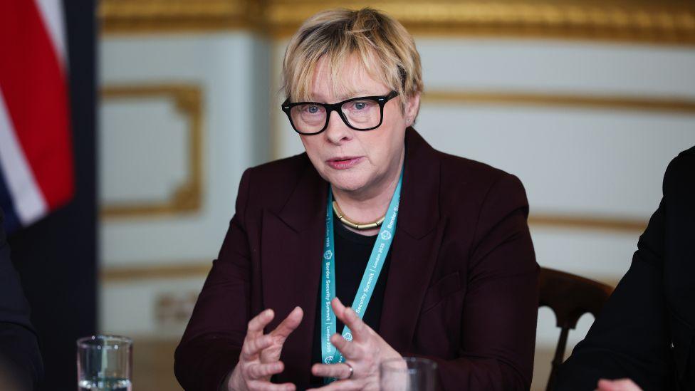 Angela Eagle, who has short blonde hair and glasses, speaks while seated at a table. She is wearing a dark suit and blue lanyard, with a union flag in the background. 