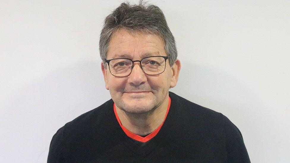 Head and shoulder shot of Alan who has short dark hair that is greying and glasses with black frames. He is smiling and is wearing a black jumper with a red T-shirt underneath. He is standing in front of a white background.