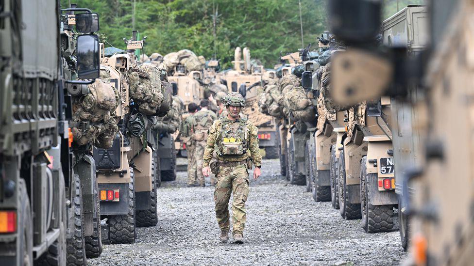 A soldier is walking towards down an avenue created by armoured vehicles either side of him. There are other colleagues in the background and they are all dressed in battle fatigues.