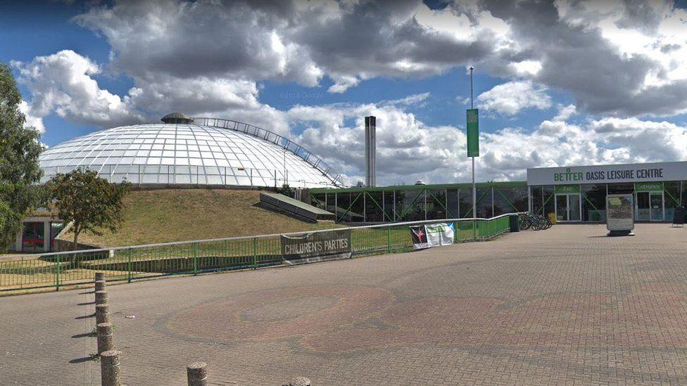 A screenshot of Google street view showing the former entrance to the Oasis leisure centre in Swindon. There is a large paved section outside the front with concrete bollards. In the distance there is the entrance lobby with double doors. On the left there is a grassy ledge leading up to the white dome where the swimming pool is.