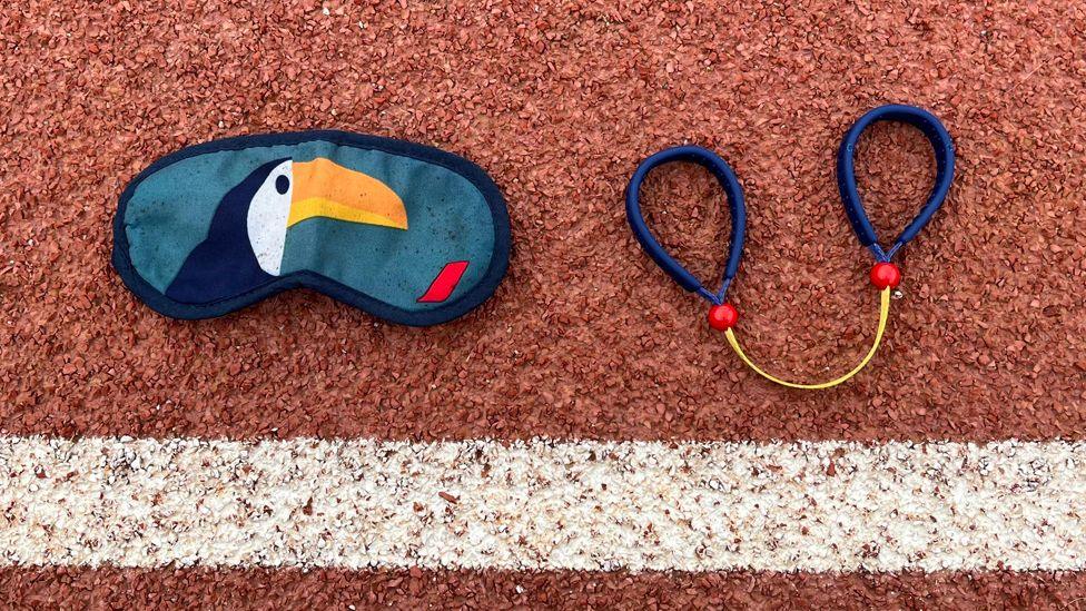 An eye mask and a tether laid on a running track. The mask has a picture of a toucan on it. The tether consists of a short yellow strap with a blue loop on each end. 